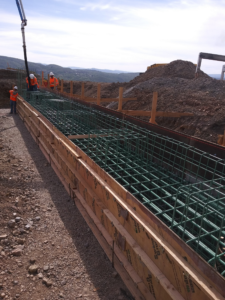 Construction workers overseeing a large concrete foundation form with green rebar at an outdoor job site.