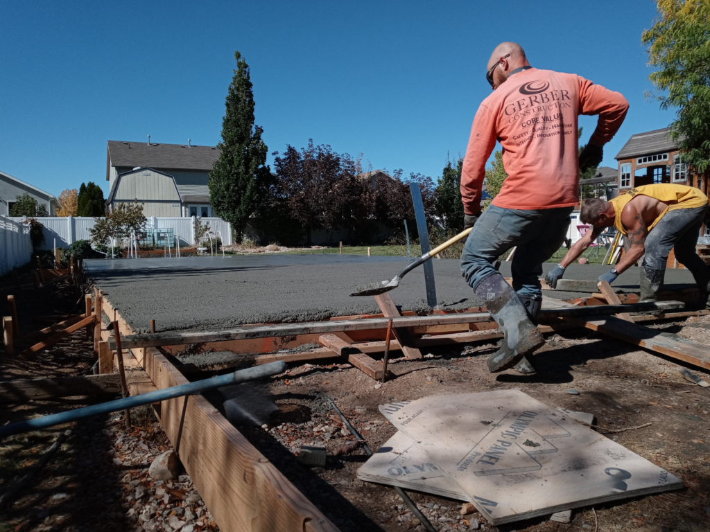 Two construction workers spreading and leveling freshly poured concrete in a residential backyard, wooden formwork lining the edges of the new concrete slab.