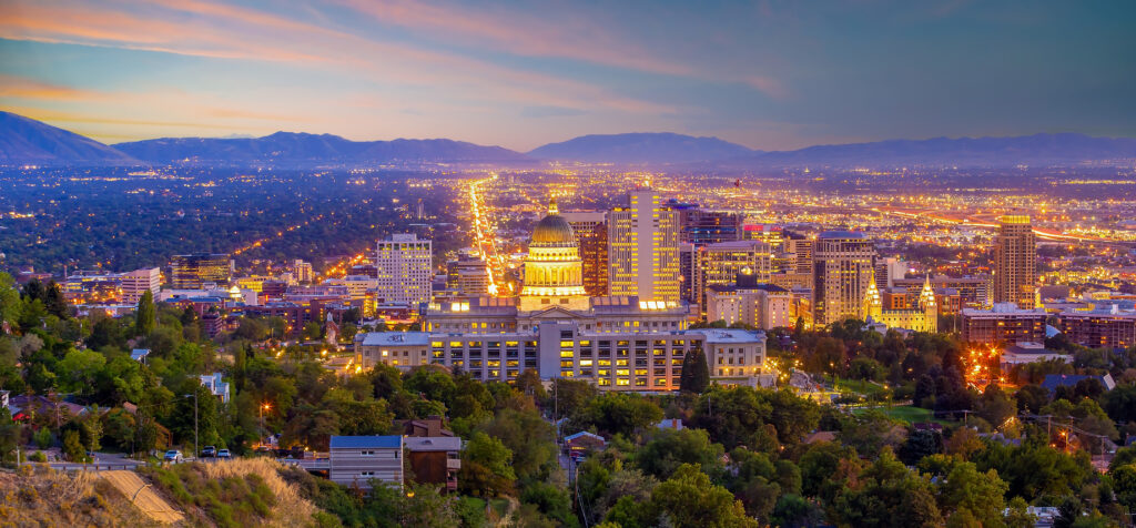 An elevated twilight view of the Salt Lake City skyline, featuring the illuminated Utah State Capitol building, glowing city lights, and distant mountains under a colorful sunset sky.