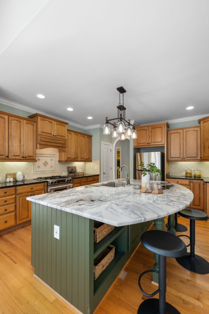 Country kitchen with sage green island, marble countertop, wood cabinetry, and a chandelier above bar seating.