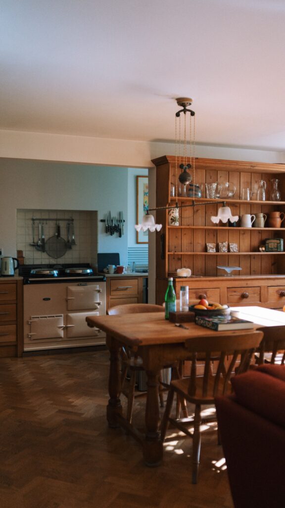 Country kitchen with cream Aga stove, wooden dining table, and a pine dresser displaying glassware and crockery.