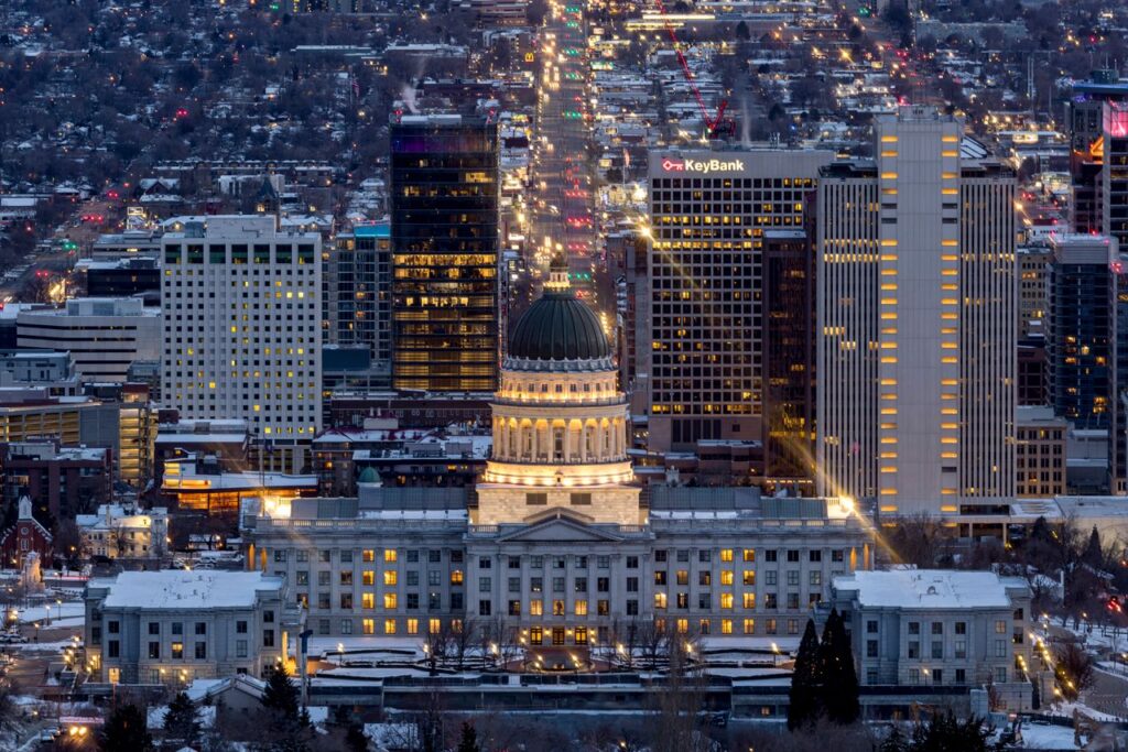 A high-angle winter night view of the illuminated Utah State Capitol, featuring snow-covered grounds and a backdrop of glowing downtown Salt Lake City skyscrapers stretching down a lit street.