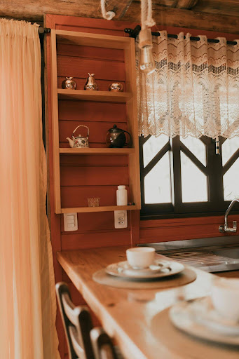 Rustic wooden kitchen nook with open shelving, vintage teapots, lace curtains, and a cozy breakfast counter.
