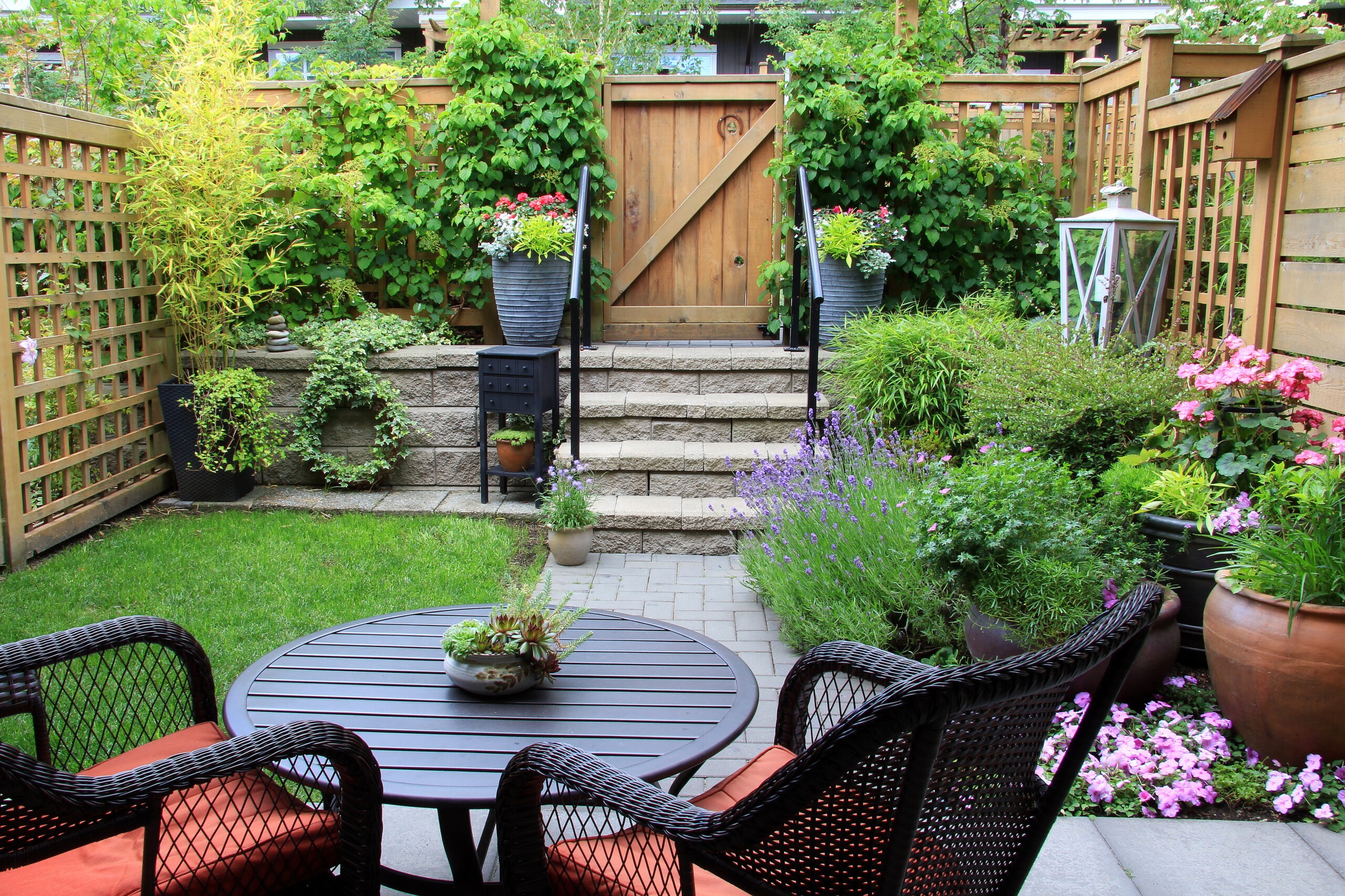 backyard garden patio with a black wicker table and chairs, stone steps leading up to a wooden gate, lush trellis greenery, and lavender and flowering plants lining a paver pathway.