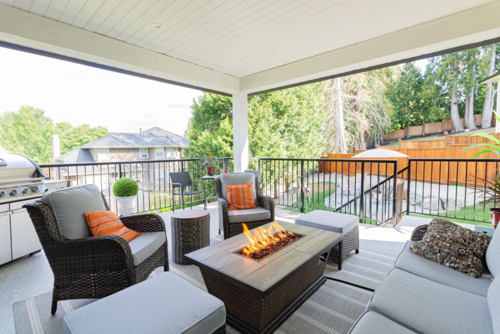Covered deck with a fire pit table, wicker seating, and a built-in outdoor grill overlooking a backyard.