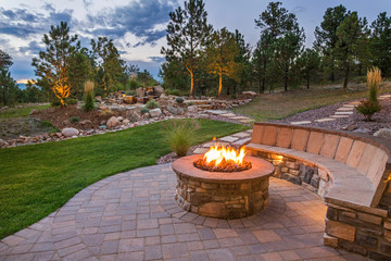 Stone patio at dusk with a round fire pit, curved stone seating wall, and a landscaped backyard with pine trees.
