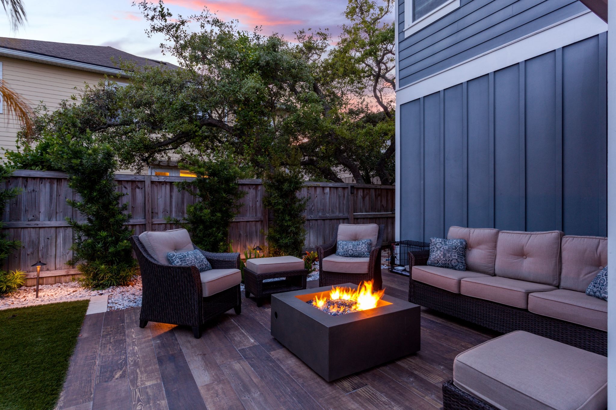 Backyard patio at dusk with a lit fire pit, wicker seating, and a sunset sky in the background.