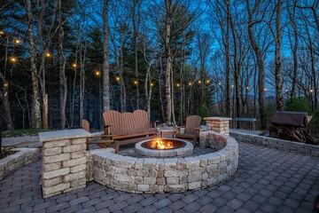 Stone patio at dusk with a circular fire pit, wooden Adirondack-style chairs, and string lights strung through bare trees.
