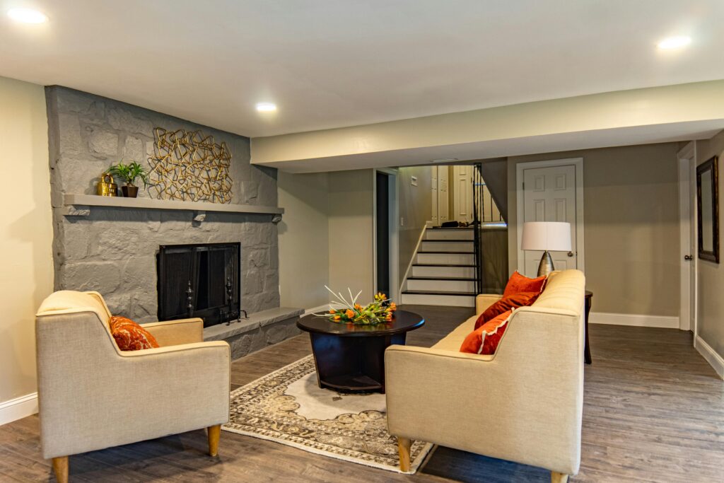 Finished basement living room with gray painted stone fireplace, cream seating, and wood plank flooring